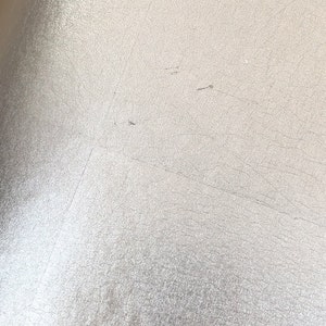 May include: Close-up of a textured, off-white surface with subtle variations in tone. The surface has a slightly metallic sheen and displays fine cracks and imperfections, creating a weathered appearance. The overall effect is a muted, neutral aesthetic.