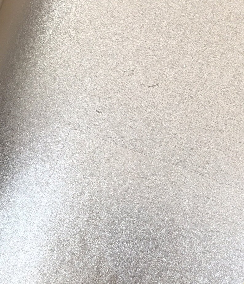 May include: Close-up of a textured, off-white surface with subtle variations in tone. The surface has a slightly metallic sheen and displays fine cracks and imperfections, creating a weathered appearance. The overall effect is a muted, neutral aesthetic.
