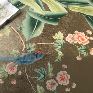 May include: Detailed illustration of a bird and floral design. The artwork features a blue bird with a red head, surrounded by peach-colored flowers and green leaves. The background has a gold-toned, textured appearance.