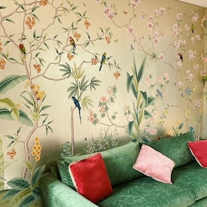 May include: A green velvet sofa with red and pink throw pillows is positioned in front of a wall covered in a botanical mural. The mural features colorful birds, flowers, and foliage in shades of green, pink, and gold.