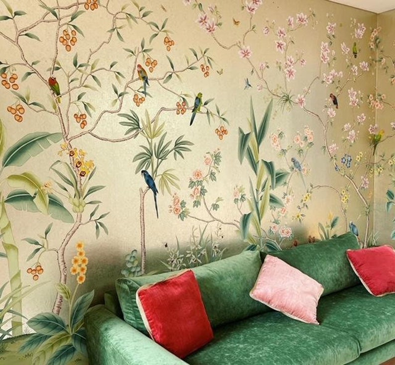 May include: A green velvet sofa with red and pink throw pillows is positioned in front of a wall covered in a botanical mural. The mural features colorful birds, flowers, and foliage in shades of green, pink, and gold.