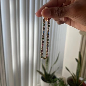May include: A gold-toned bracelet with a series of colorful, rectangular and oval-shaped gemstones. The gemstones are in shades of blue, green, red, and purple. The bracelet is held up against a white curtain and potted plants.