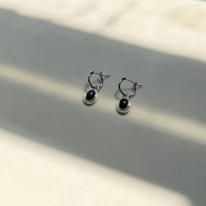 May include: A pair of silver hoop earrings with oval, black gemstones. The earrings are displayed on a white surface, with shadows creating a soft contrast. The jewelry is simple and elegant.