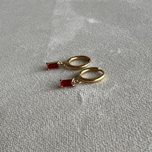 May include: A pair of gold-colored hoop earrings with rectangular, ruby-red gemstones dangling from the hoops. The earrings are displayed on a textured, off-white surface. The earrings are a jewelry item.