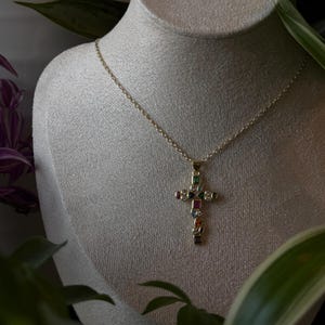 May include: A gold-colored cross pendant necklace with colorful gemstones. The cross is displayed on a mannequin, with green foliage in the background. The gemstones are various colors, including red, blue, and green.