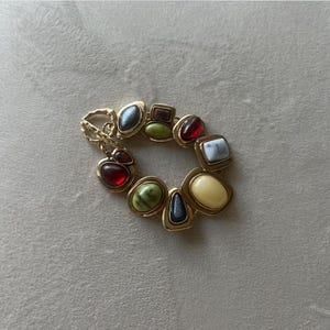 May include: A gold-toned bracelet featuring a variety of colorful, irregularly shaped gemstones. The gemstones include red, green, blue, and white stones, set in a gold metal frame. The bracelet is a statement piece.