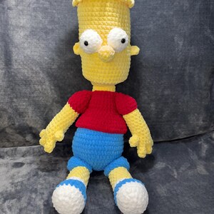May include: A crocheted plush toy of Bart Simpson from The Simpsons. The toy is yellow with a red shirt, blue shorts, and white shoes.