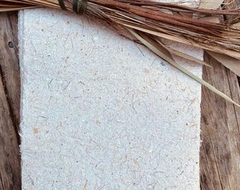 Può includere: Una pila di carta fatta a mano con bordo a risvolto, con fibre vegetali e scaglie visibili. Un fascio di erbe e foglie secche, legato con una fascia verde scuro, è appoggiato sopra. La carta è di colore bianco sporco naturale.