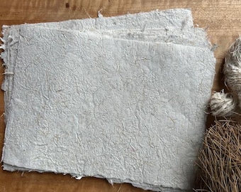 Può includere: Una pila di carta fatta a mano, con una superficie strutturata e bordi irregolari. La carta è grigio chiaro con fibre visibili. Un fascio di spago e materiale vegetale secco sono sullo sfondo.