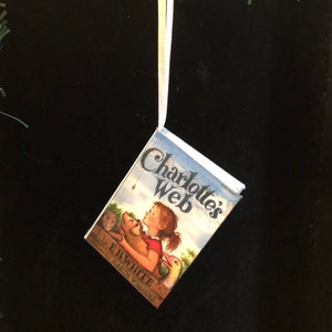 May include: A miniature book ornament featuring the cover of Charlotte's Web by E.B. White. The cover is blue and white with illustrations of a pig, a spider, and a girl. The ornament is hanging from a white string.
