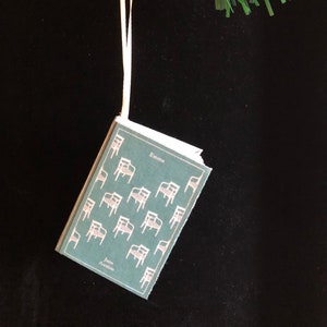 May include: A green book ornament with a white ribbon hanging from a green Christmas tree branch. The book cover features a repeating pattern of chairs and the text "Emma" and "Jane Austen".