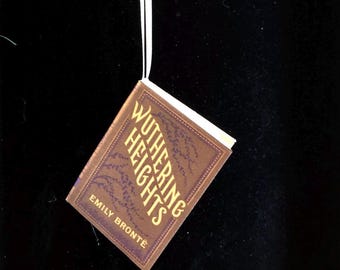 May include: A miniature book ornament titled Wuthering Heights by Emily Brontë, featuring a brown cover with gold lettering and dark purple botanical illustrations, suspended by a thin white ribbon for hanging as a decorative item.