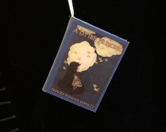 May include: A blue book with the title "A Little Princess" and the author's name "Frances Hodgson Burnett" printed on the cover. The cover features an illustration of a young girl standing on a cloud with birds flying around her.