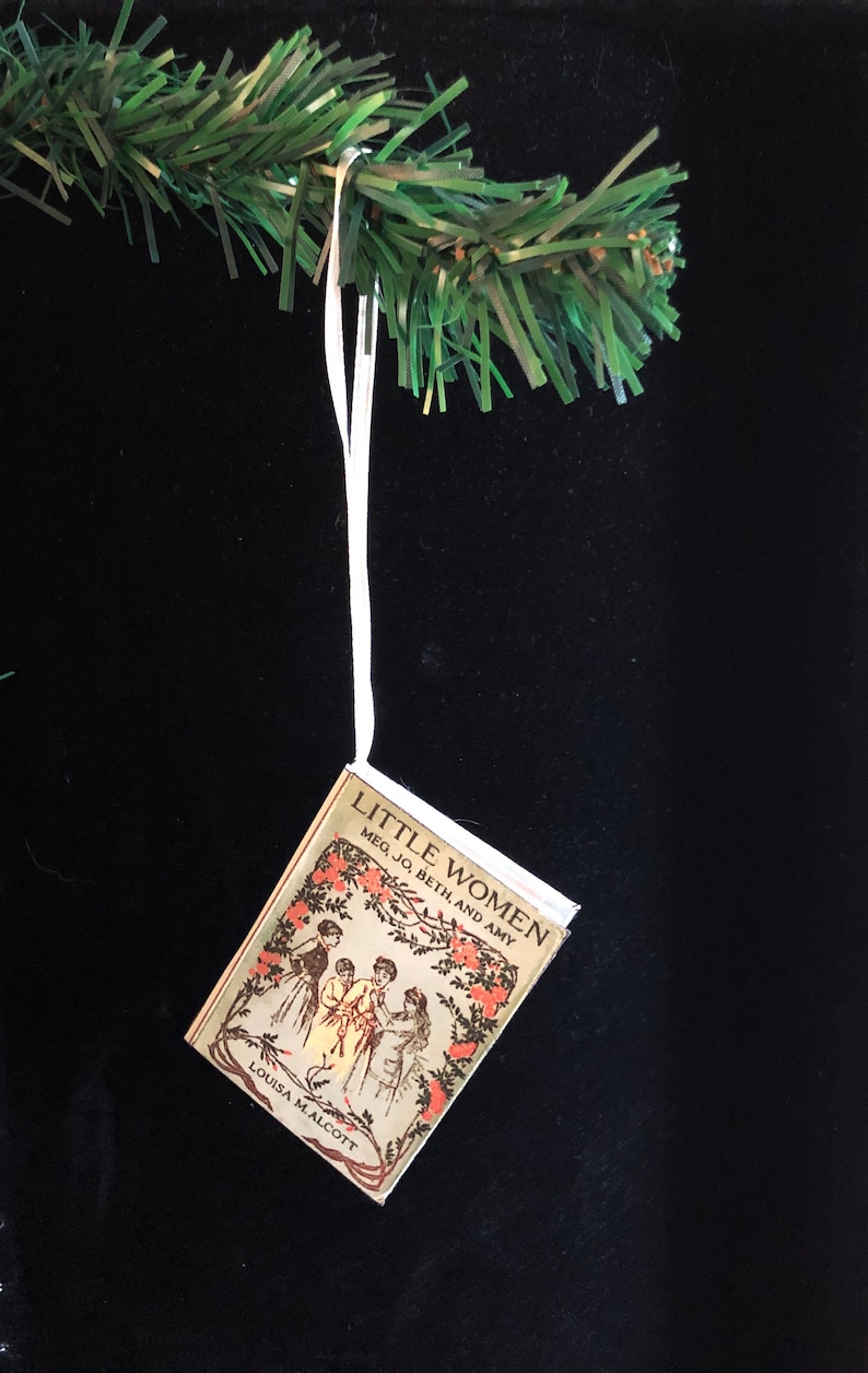 May include: A miniature book ornament featuring the cover of "Little Women" by Louisa May Alcott. The cover is illustrated with four girls in a garden setting. The book is hanging from a white string.
