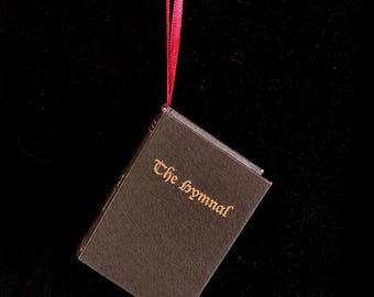 May include: A miniature brown book ornament titled The Hymnal in gold lettering, suspended by a thin red ribbon. The cover has a textured surface and the spine features gold lines. The book hangs at an angle against a dark background.