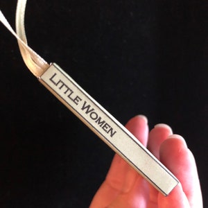 May include: A close-up of a beige bookmark with the text "LITTLE WOMEN" printed in black.