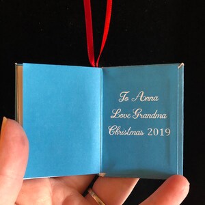 May include: A small blue book ornament with a red ribbon. The book is open and the inside reads "To Anna Love Grandma Christmas 2019".