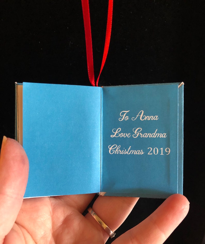 May include: A small blue book ornament with a red ribbon. The book is open and the inside reads "To Anna Love Grandma Christmas 2019".
