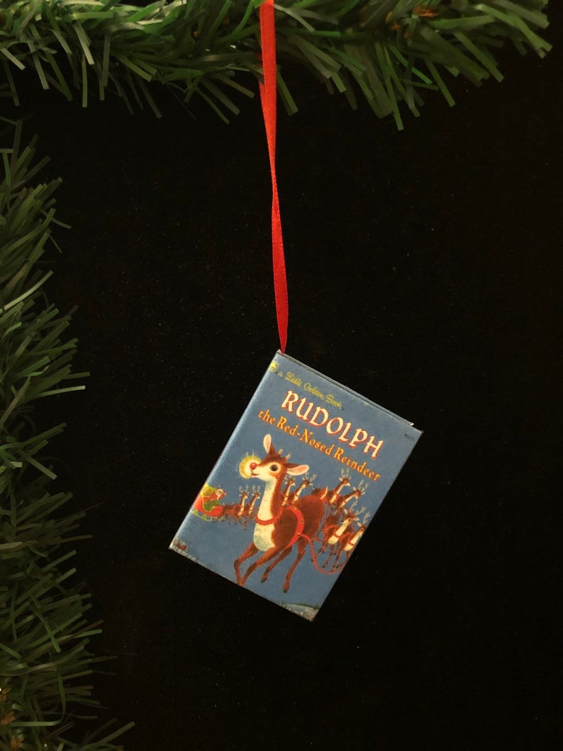 May include: A miniature book ornament featuring Rudolph the Red-Nosed Reindeer. The book is blue with a red ribbon attached. The cover features a colourful illustration of Rudolph leading a sleigh.