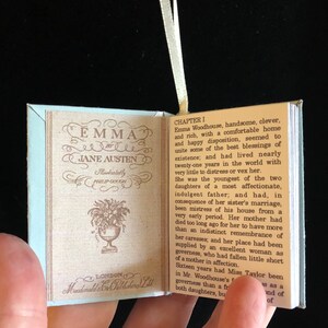 May include: A miniature book replica of Jane Austen's novel "Emma" with a light blue cover and a white ribbon bookmark. The book is open to the first chapter and features a floral illustration on the left page.