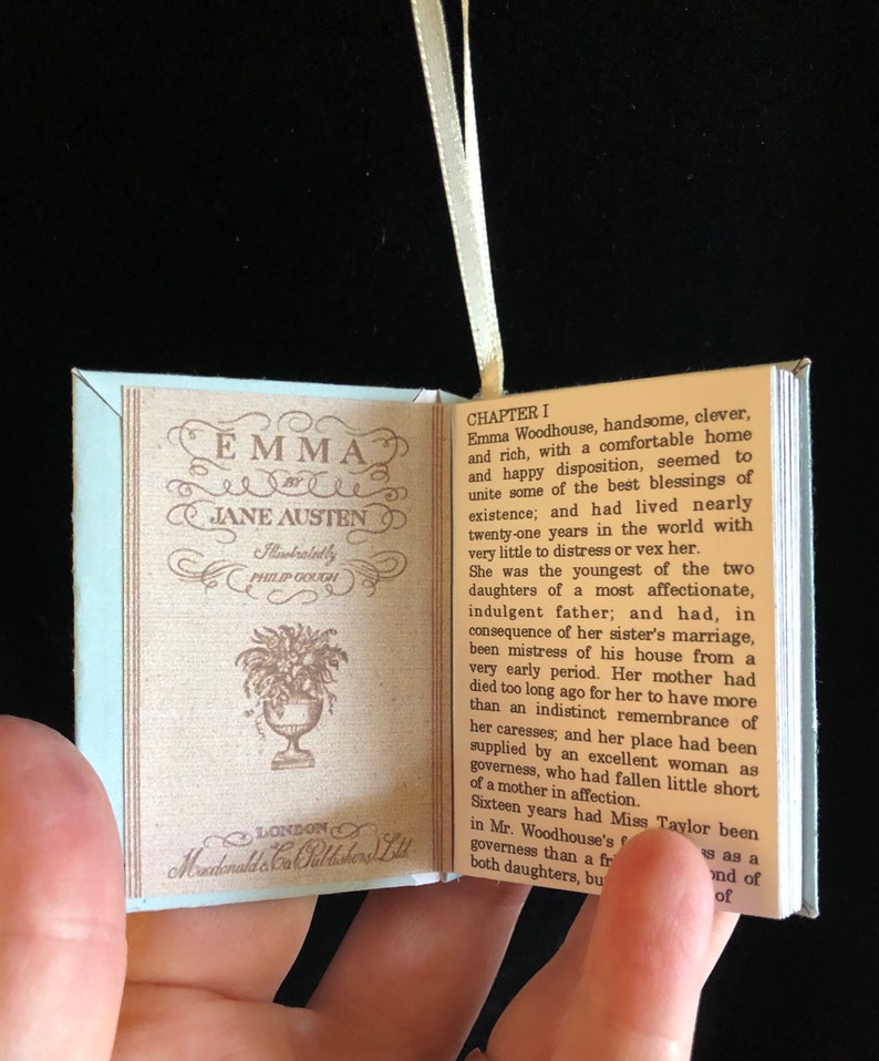 May include: A miniature book replica of Jane Austen's novel "Emma" with a light blue cover and a white ribbon bookmark. The book is open to the first chapter and features a floral illustration on the left page.