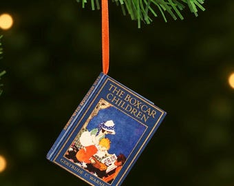May include: A miniature book ornament titled The Box-Car Children by Gertrude C. Warner, featuring a blue cover with a vintage illustration of children reading, suspended by a thin orange ribbon from a green artificial pine branch.
