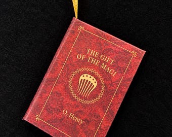 May include: A miniature book titled The Gift of the Magi by O. Henry with a red cover featuring a gold decorative pattern and a central gold comb icon. The book is suspended by a thin yellow ribbon attached to the top edge of the cover.