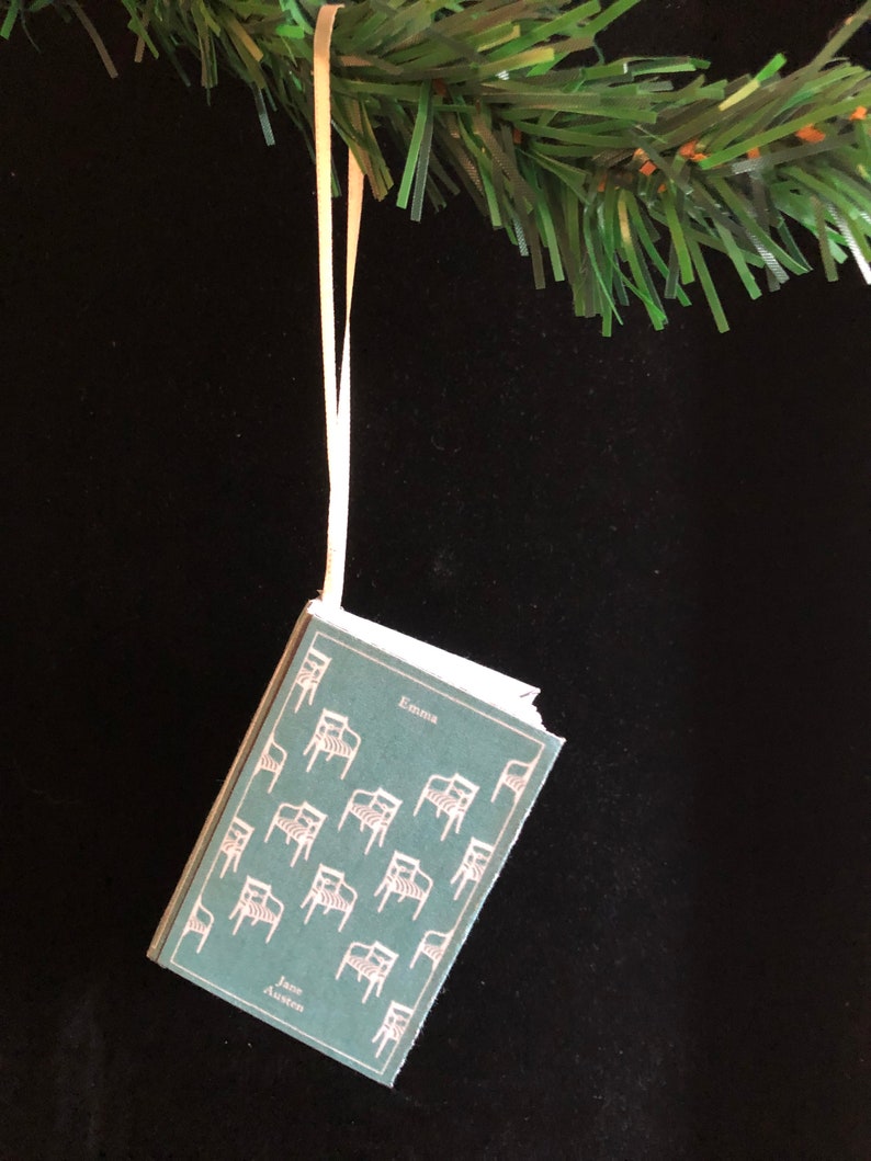 May include: A green book ornament with a white ribbon hanging from a green Christmas tree branch. The book cover features a repeating pattern of chairs and the text "Emma" and "Jane Austen".