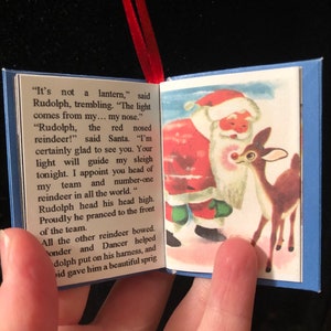 May include: A vintage book ornament featuring Rudolph the Red-Nosed Reindeer. The book is open to a page with an illustration of Santa Claus and Rudolph. The text reads, "It's not a lantern," said Rudolph, trembling. "The light comes from my... my nose."  "Rudolph, the red nosed reindeer!" said Santa. "I'm certainly glad to see you. Your light will guide my sleigh tonight. I appoint you head of my team and number-one reindeer in all the world."