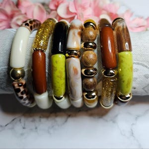Peut inclure: Un empilement de sept bracelets composés de perles variées, incluant des cylindres lisses crème, marron et vert citron, des accents dorés pailletés, des tubes noirs polis, des pièces marbrées blanc et marron, et des sphères en bois naturel.