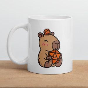 Puede incluir: Taza de cerámica blanca con una capibara de dibujos animados que lleva un pequeño dado de 20 caras naranja en la cabeza y sostiene un dado de 20 caras naranja más grande en sus patas.