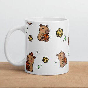 Puede incluir: Taza de cerámica blanca con un patrón repetido de capibaras marrones que sostienen dados naranjas y estrellas verdes de cuatro puntas.