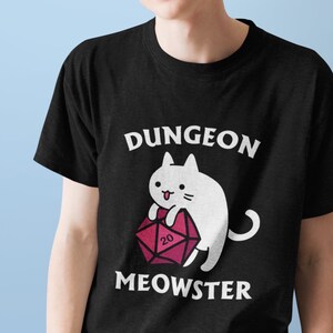 以下が含まれることがあります： 白い猫が赤い20面ダイスを持っている黒いTシャツ。Tシャツには「Dungeon Meowster」という文字が印刷されています。