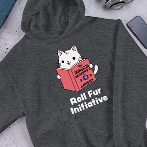 May include: Dark gray hoodie featuring a white cat reading a red book titled "The Dungeon Meowster's Handbook." The text "Roll Fur Initiative" is printed below the cat. The hoodie is displayed with blue jeans, headphones, and other accessories.