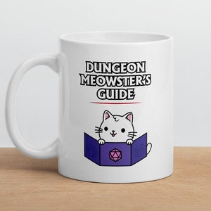 May include: White ceramic mug with a black and red graphic that says "Dungeon Meowster's Guide". A white cat with pink cheeks is holding a purple book with a pink and white dice inside.