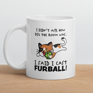 May include: White ceramic mug featuring a cartoon cat with a green die, and the text "I DIDN'T ASK HOW BIG THE ROOM WAS. I SAID I CAST FURBALL!". The cat is black, orange, and white.