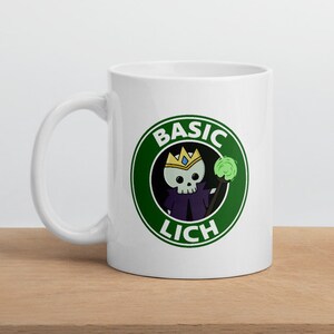 May include: White ceramic mug with a green circle design featuring a cartoon skeleton wearing a crown and holding a staff. The text "BASIC LICH" is written in the circle.