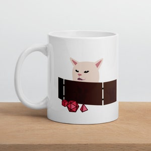 May include: White ceramic mug with a white cat illustration inside a brown dungeon with red dice. The cat has a grumpy expression.