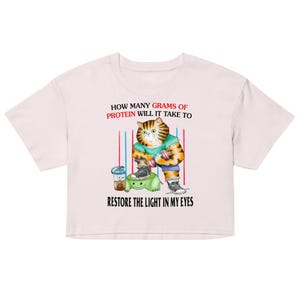 Könnte beinhalten: Ein hellrosa Crop-Top mit einer Cartoon-Katze, die Gewichte hebt. Die Katze trägt ein gestreiftes Hemd und Shorts. Auf dem Shirt steht: "HOW MANY GRAMS OF PROTEIN WILL IT TAKE TO RESTORE THE LIGHT IN MY EYES."