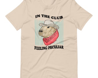 May include: A beige t-shirt with a cartoon capybara wearing a cowboy hat and bandana. The text "IN THE CLUB FEELING PECULIAR" is printed on the shirt.