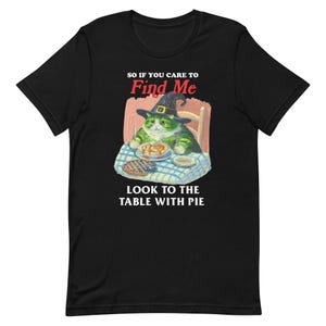 Puede incluir: Camiseta negra con un gráfico blanco de un gato con un sombrero de bruja y comiendo pastel en una mesa. El texto dice "So if you care to Find Me Look to the table with pie."
