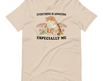 Puede incluir: Una camiseta beige con un gato de dibujos animados sentado debajo de un hongo. El texto "Todo es molesto, especialmente yo" está impreso en la camiseta.
