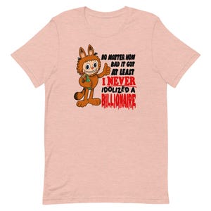 Könnte beinhalten: Hellrosa T-Shirt mit einer Comicfigur, die den Daumen hochhält. Die Figur hält eine Flasche, und der Text lautet: "NO MATTER HOW BAD IT GOT AT LEAST I NEVER IDOLIZED A BILLIONAIRE."