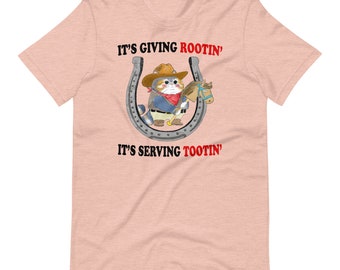 May include: A light pink t-shirt featuring a cartoon cat dressed as a cowboy inside a horseshoe. The cat wears a hat, red bandana, and holds a toy horse. Text on the shirt reads "IT'S GIVING ROOTIN'" and "IT'S SERVING TOOTIN'" in red.