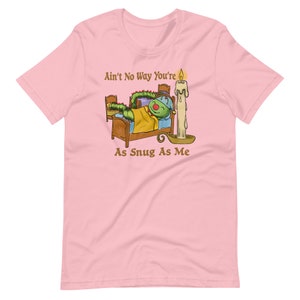 Könnte beinhalten: Pinkes T-Shirt mit einer Cartoon-Illustration einer grünen Raupe im Bett mit einer Kerze. Der Text auf dem Shirt lautet "Ain't No Way You're As Snug As Me."