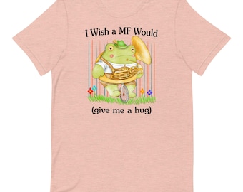 Puede incluir: Una camiseta rosa claro con una rana verde que lleva un sombrero y toca una tuba. La rana está montada en un monociclo. El texto de la camiseta dice "I Wish a MF Would (give me a hug)"