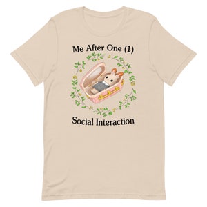 May include: A beige t-shirt featuring a graphic of a bunny in a pink coffin surrounded by green leaves and yellow flowers. The text "Me After One (1) Social Interaction" is printed on the shirt.