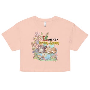 May include: A pink cropped t-shirt with a graphic of two cats sleeping in a pond surrounded by flowers and mushrooms. The text "Lowkey Plotting & Scheming" is above the cats.