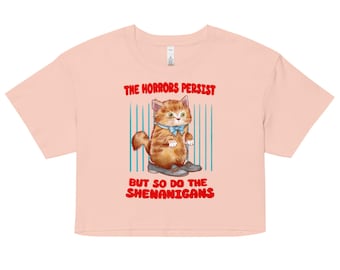 May include: A pale pink crop top with a cartoon cat wearing shoes and a blue bow tie. The text "THE HORRORS PERSIST" and "BUT SO DO THE SHENANIGANS" are printed in red. The cat is set against a background of blue vertical lines.