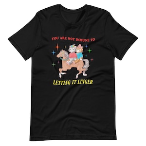 May include: Black t-shirt featuring a graphic of two cats riding a horse. The text "YOU ARE NOT IMMUNE TO LETTING IT LINGER" is printed in red and yellow.